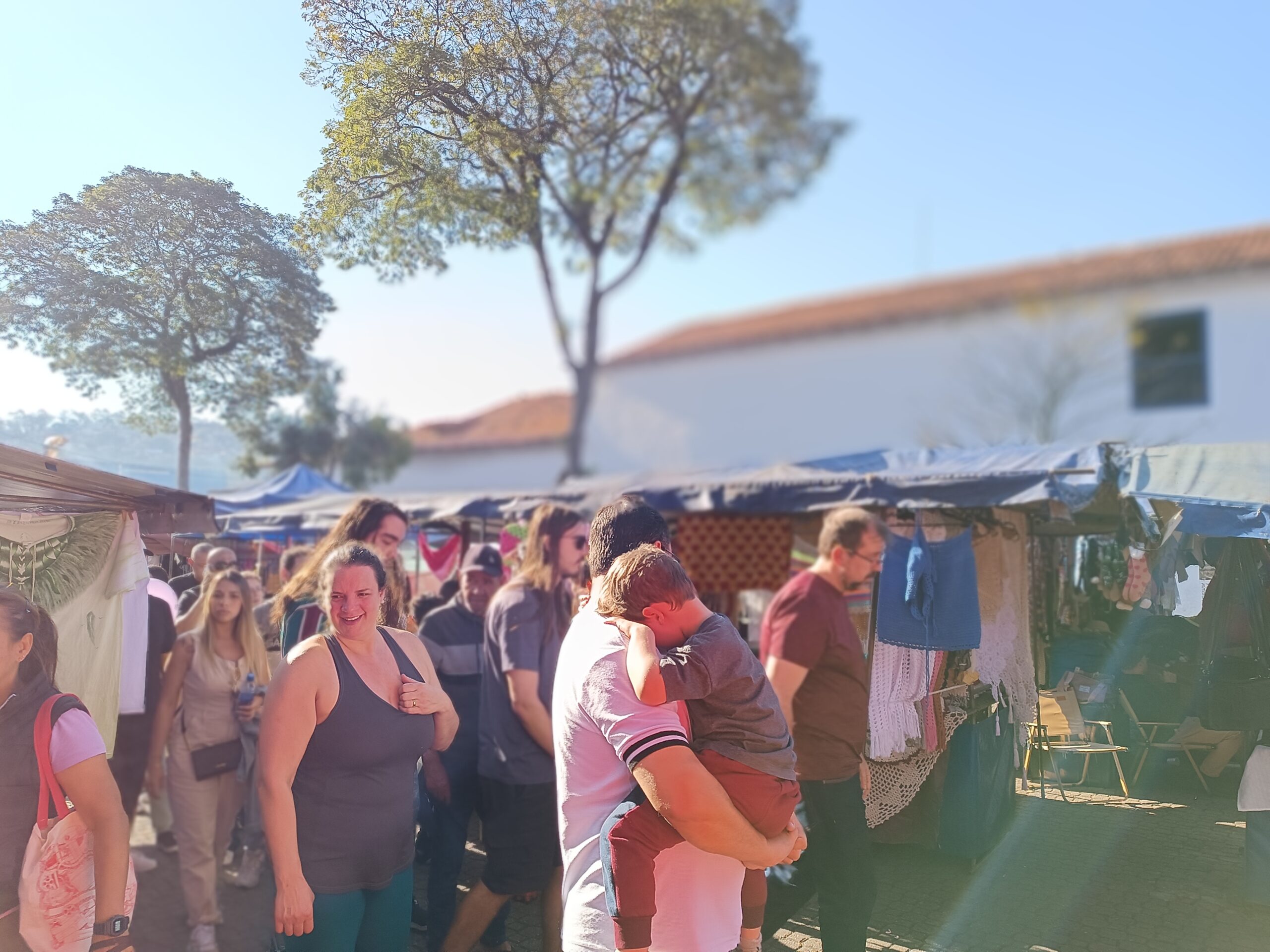 Visitantes circulando pelo Largo dos Jesuítas, ao lado do Museu de Arte Sacra de Embu das Artes, observando os diversos espaços da tradicional Feira de Embu das Artes e os trabalhos apresentados pelos expositores.